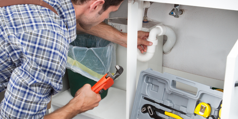 Plumber holding a straight wrench and working under a kitchen sink.