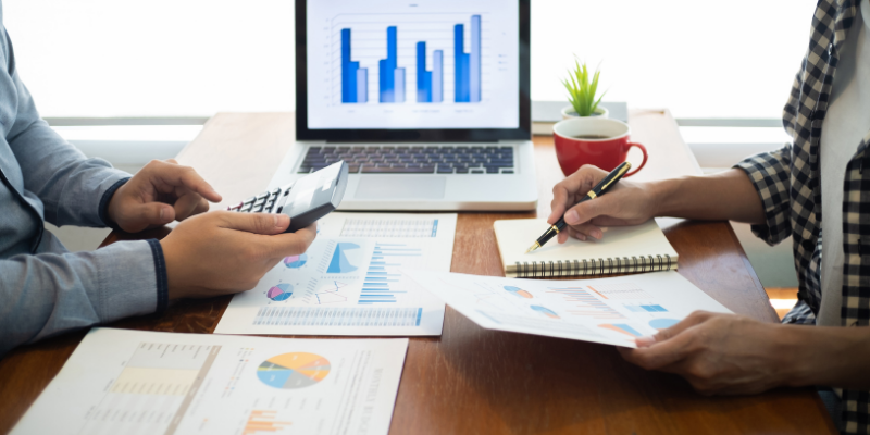 Two people sharing a table with financial statements laid out, a calculator, and a computer. 