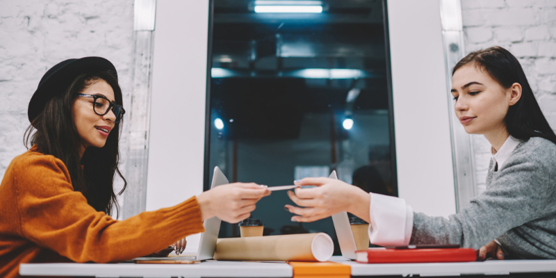 Two women sitting at a table and exchanging a piece of paper